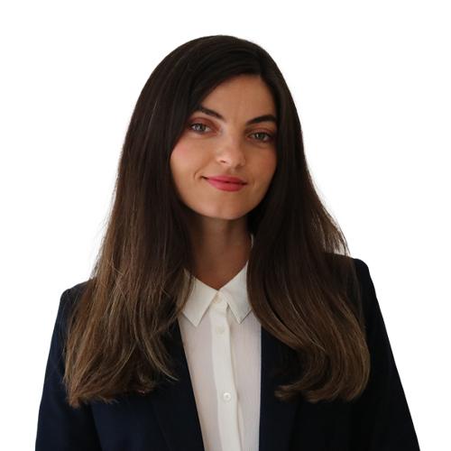 Professional headshot of a woman with long dark hair, wearing a white collared shirt and navy blazer, smiling slightly.